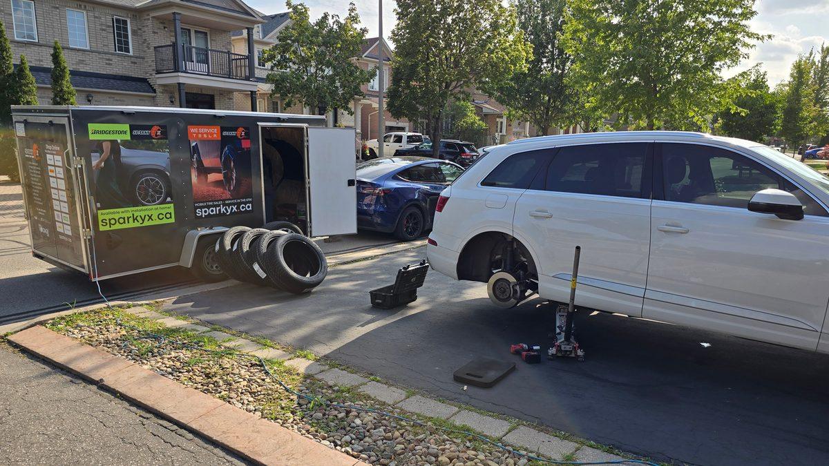 Sparky X mobile tire services at work — a white Audi Q7 on a jack in a driveway, rear wheel off, the Sparky X trailer open beside it with tires stacked out and full trailer branding visible