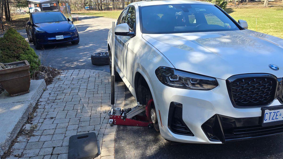 European car tire service by Sparky X — a white BMW X4 on a red jack in a paved driveway, front wheel off, the blue Sparky X Model Y and service trailer behind