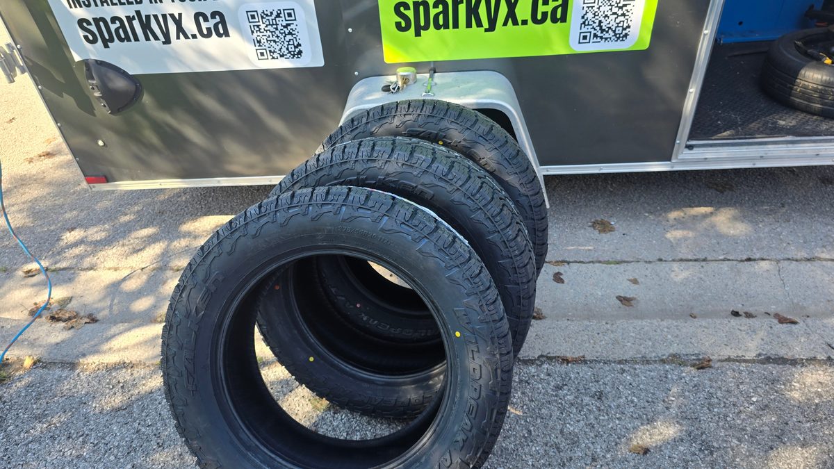 Three new Falken Wildpeak A/T tires leaning against the Sparky X trailer — sparkyx.ca signage in frame, ready for free driveway installation