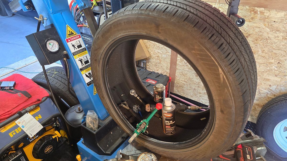 Flat tire repair by Sparky X inside the mobile trailer — a tire mounted on the blue changer mid repair, repair supplies and tools in its centre, the generator at the left