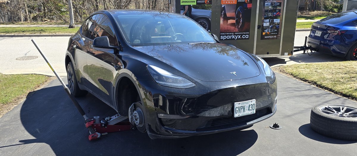Black Tesla SUV on a jack in a Bowmanville driveway during a Sparky X mobile tire service — trailer behind, mounted wheels in the foreground