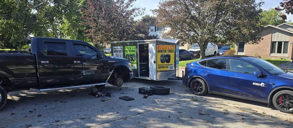 Black pickup truck on a jack during a Sparky X mobile tire service in Pickering — a blue Tesla and the Sparky X trailer behind