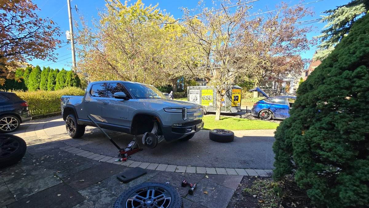 Sparky X EV tire service at work — a grey Rivian R1T on a jack in an upscale driveway, front wheel off with the removed wheel and tools on the stone pavers, the Sparky X service trailer and a blue Tesla behind it