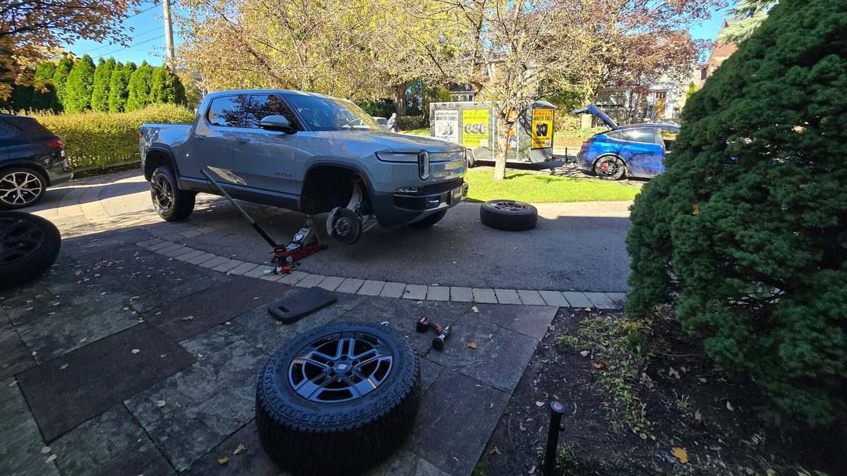 Sparky X seasonal tire changeover in a customer's driveway — a Rivian R1T on a jack with its front wheel off, a Pirelli Scorpion tire and wheel ready in the foreground, the Sparky X trailer and a Tesla Model Y behind