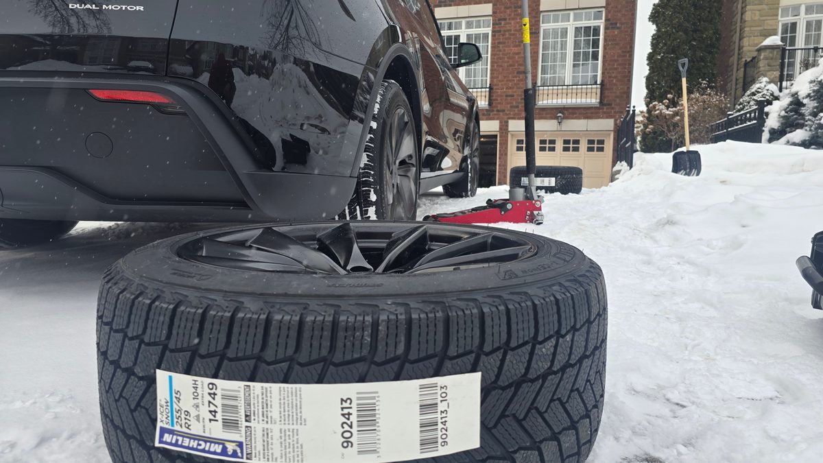 Sparky X winter tire package in a snowy driveway — a new Michelin X-Ice Snow 255/45R19 on a black wheel in front of a black Tesla Model Y, red floor jack behind, ready for free mobile installation