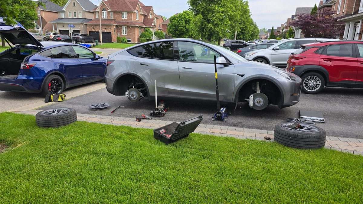 Tire rotation by Sparky X at a customer's home — a grey Tesla Model Y on two jacks in a residential driveway with all four wheels off, the wheels and tools laid out around it, the blue Sparky X Model Y parked behind