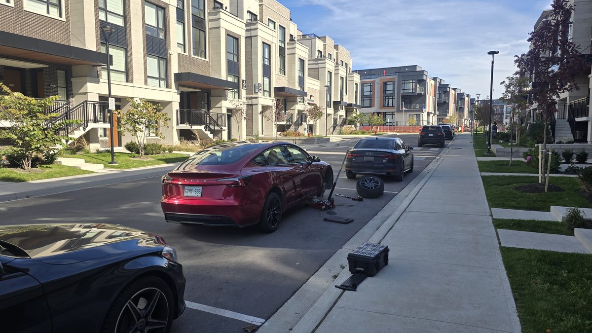 Red Tesla Model 3 on a jack mid tire change on a residential street — front wheel off, the removed wheel and tools on the pavement beside it, Sparky X mobile tire service at work